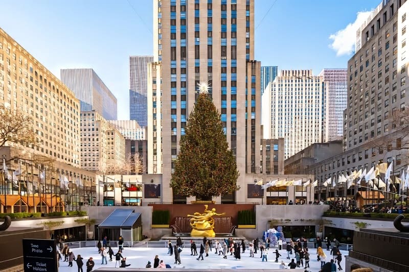 Các vận động viên trượt băng lướt nhẹ trên sân băng bên dưới một cây thông Noel cao vút được trang trí lộng lẫy tại Trung tâm Rockefeller. Những tòa nhà chọc trời vươn cao phía sau, gợi lên một không khí lễ hội.