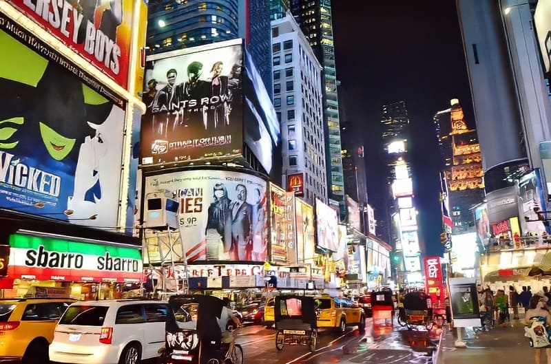 Quảng trường Times Square về đêm, nhộn nhịp với những chiếc taxi màu vàng và người đi bộ. Những biển quảng cáo rực rỡ chiếu sáng đường phố, tạo nên một bầu không khí năng động và sống động.