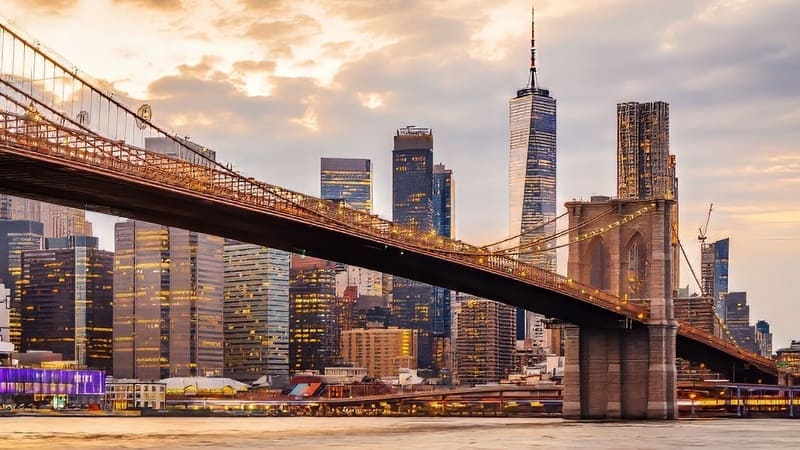 Cảnh cầu Brooklyn lúc hoàng hôn với đường chân trời rực rỡ của thành phố New York phía sau, nổi bật với những tòa nhà chọc trời dưới bầu trời nhiều mây đầy màu sắc.