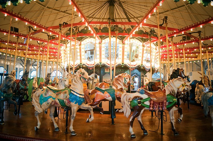 Một chiếc đu quay ngựa gỗ sáng rực rỡ với những chú ngựa được trang trí công phu và đầy màu sắc được đặt trong nhà. Ánh sáng ấm áp và những đồ trang trí tinh xảo tạo nên một bầu không khí hoài niệm và vui tươi.