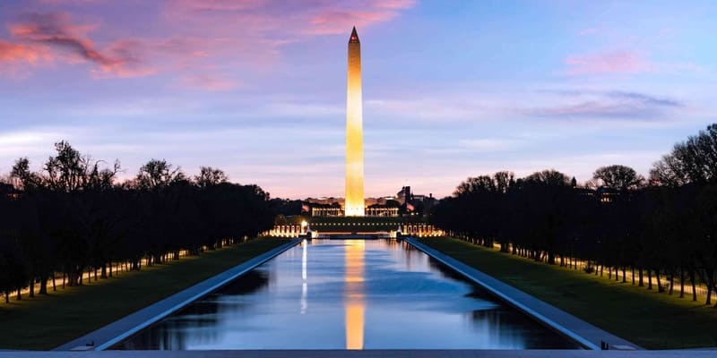 Cột đá obelisk cao vút, Đài tưởng niệm Washington, phản chiếu trong một hồ nước dài lúc hoàng hôn. Bầu trời rực rỡ sắc hồng và xanh lam, bao quanh là những hàng cây in bóng.