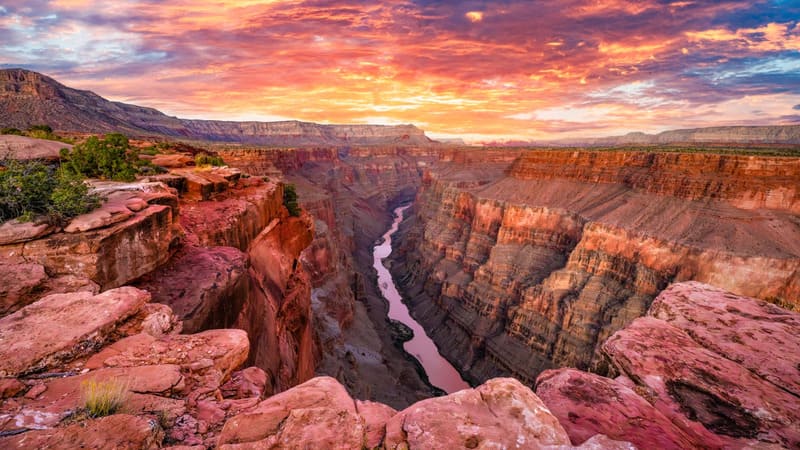 Hoàng hôn rực rỡ chiếu sáng Grand Canyon, làm nổi bật những lớp đá đỏ xếp chồng lên nhau và hẻm núi sâu hun hút. Bầu trời rực cháy với những sắc hồng và cam.