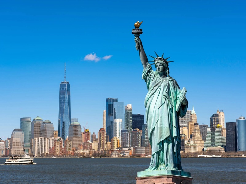 Tượng Nữ thần Tự do sừng sững ở tiền cảnh, phía sau là đường chân trời thành phố New York và bầu trời xanh trong vắt. Một chiếc phà đang băng qua mặt nước.