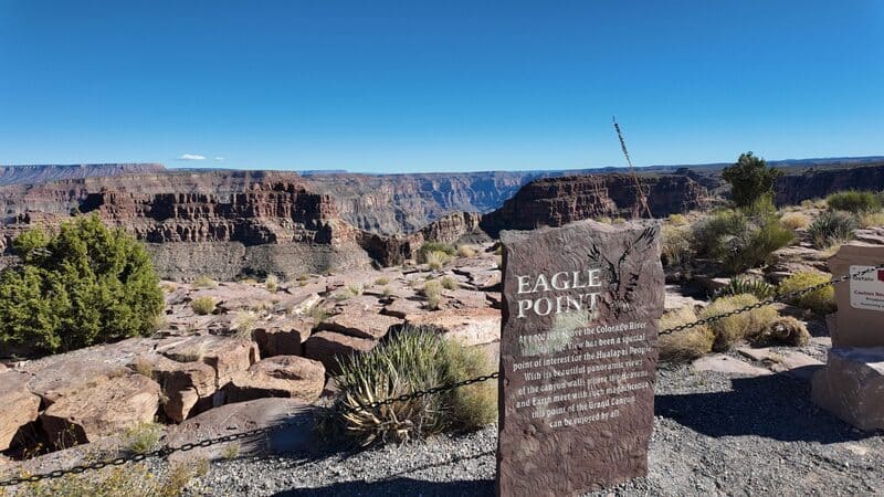 Điểm Eagle Point tại Grand Canyon; những vách đá hiểm trở dưới bầu trời xanh trong vắt. Một tấm biển thông tin nằm ở phía trước giữa thảm thực vật sa mạc thưa thớt.