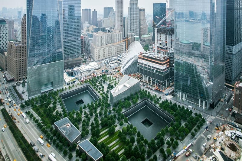 Ảnh chụp từ trên cao Đài tưởng niệm 11/9 của thành phố New York, với hai hồ nước lớn phản chiếu ánh sáng được bao quanh bởi cây cối. Những tòa nhà chọc trời và những con phố nhộn nhịp tạo nên khung cảnh tuyệt đẹp.