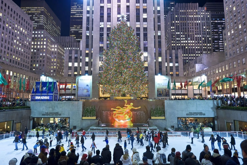 Một cây thông Noel rực rỡ được trang trí bằng đèn màu sắc nổi bật sừng sững tại Trung tâm Rockefeller, bao quanh là những tòa nhà chọc trời. Những người trượt băng đang tận hưởng sân băng bên dưới trong không khí lễ hội.