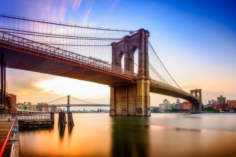 Cầu Brooklyn bắc qua mặt nước tĩnh lặng lúc hoàng hôn với bầu trời đầy màu sắc. Đường chân trời thành phố hiện lên ở phía xa, tạo nên một khung cảnh đô thị thanh bình.
