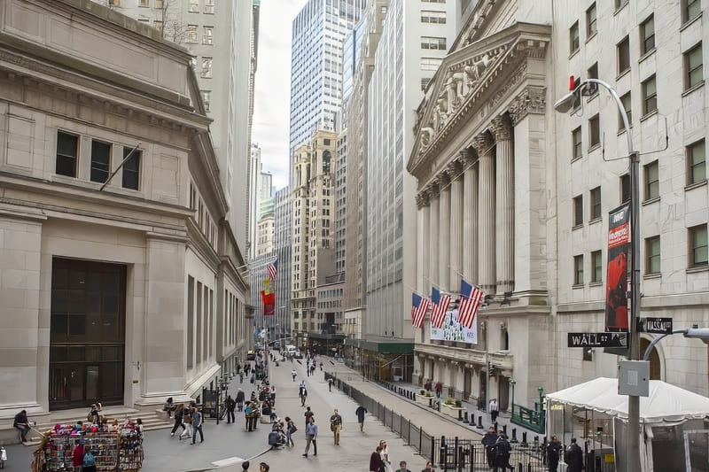 Khung cảnh nhộn nhịp của phố Wall với người đi bộ ngang qua Sở Giao dịch Chứng khoán New York. Cờ Mỹ được treo trên tòa nhà, thể hiện năng lượng và tầm quan trọng của nó.