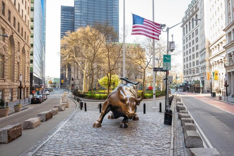 Tượng bò tót bằng đồng trên con phố lát đá cuội ở khu tài chính của thành phố New York, với quốc kỳ Mỹ và các tòa nhà lịch sử ở phía sau.