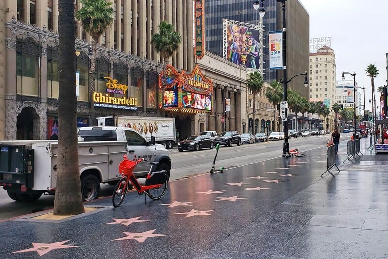 Hình ảnh cho thấy Đại lộ Hollywood, nổi bật với Con đường Danh vọng được gắn những ngôi sao. Một chiếc xe đạp và xe tay ga nằm rải rác trên vỉa hè ướt.