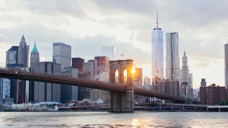 Cảnh hoàng hôn nhìn ra đường chân trời thành phố New York với cầu Brooklyn ở phía trước. Ánh nắng len lỏi qua các vòm cầu, chiếu rọi ánh sáng ấm áp lên những tòa nhà cao tầng.