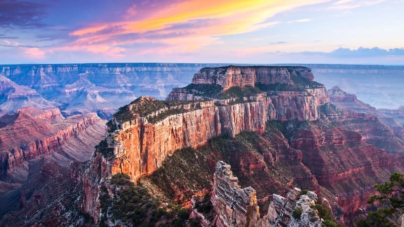 Cảnh hoàng hôn rực rỡ trên Grand Canyon, làm nổi bật những vách đá màu đỏ và cam xếp lớp. Sắc tím và hồng của bầu trời càng làm tăng thêm vẻ thanh bình, hùng vĩ.