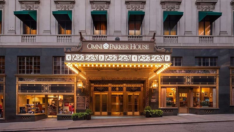 Lối vào khách sạn trang nhã về đêm, mang biển hiệu "Omni Parker House". Ánh đèn ấm áp chiếu sáng mặt tiền lộng lẫy, với những chi tiết trang trí cầu kỳ và cây cảnh trong chậu. Cổ điển và mời gọi.