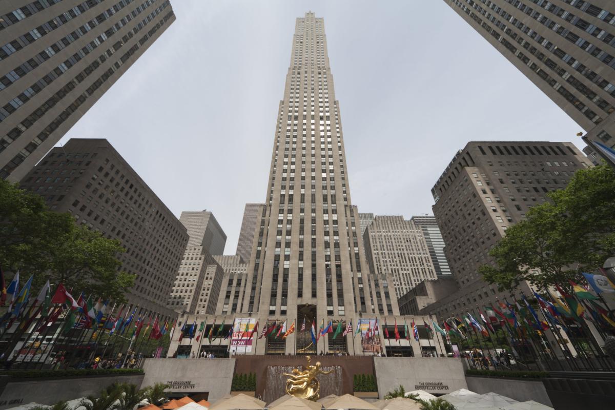 Tòa nhà chọc trời cao vút tại Rockefeller Center, New York, với bức tượng vàng phía trước và những lá cờ quốc tế xếp dọc quảng trường, thể hiện sự uy nghi và đoàn kết toàn cầu.
