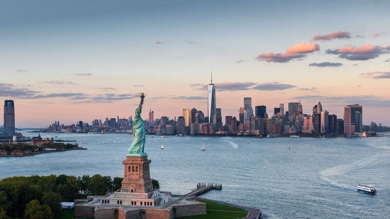 Hình ảnh từ trên cao của Tượng Nữ thần Tự do với đường chân trời thành phố New York ở phía sau lúc hoàng hôn. Khung cảnh yên bình với mặt nước tĩnh lặng.
