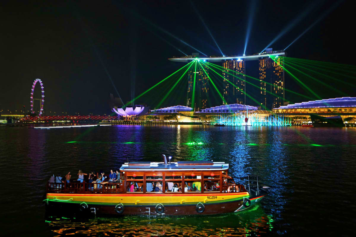 Tour singapore 4 ngày 3 đêm: Một khung cảnh buổi tối sống động tại Vịnh Marina của Singapore, với ánh đèn laser đầy màu sắc từ Marina Bay Sands, một chiếc thuyền ở phía trước và một vòng đu quay.