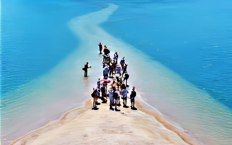 Một nhóm người tụ tập trên một dải cát hẹp bao quanh bởi làn nước xanh thẳm dưới bầu trời trong xanh. Khung cảnh vui tươi này truyền tải cảm giác phiêu lưu và khám phá.