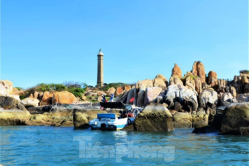 Một chiếc thuyền neo đậu gần bờ đá, phía sau là ngọn hải đăng cao vút dưới bầu trời xanh trong vắt.