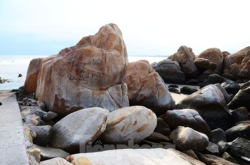 Những tảng đá lớn ven biển có khắc chữ gần đại dương, được bao quanh bởi những viên đá nhỏ hơn dưới bầu trời trong xanh, tạo nên một khung cảnh thanh bình.