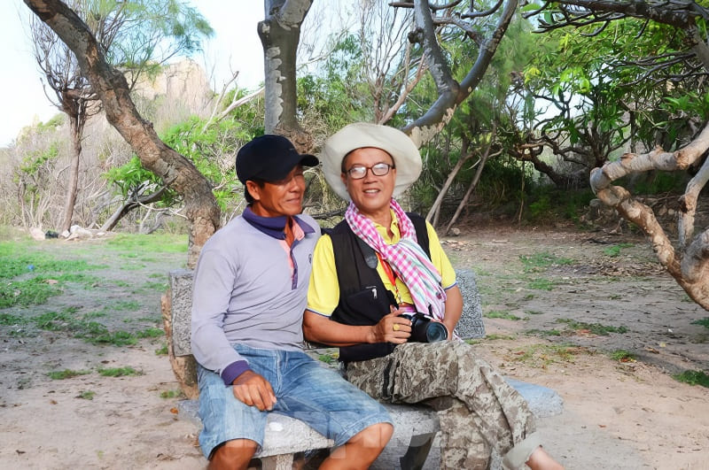 Hai người đàn ông ngồi trên ghế đá công viên, một người cầm máy ảnh. Xung quanh họ là cây cối và thảm thực vật xanh tươi, tận hưởng một khoảnh khắc yên bình.