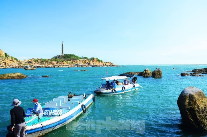 Những chiếc thuyền lướt nhẹ trên mặt biển màu ngọc lam gần bờ đá, phía xa là ngọn hải đăng trên ngọn đồi dưới bầu trời xanh trong vắt.