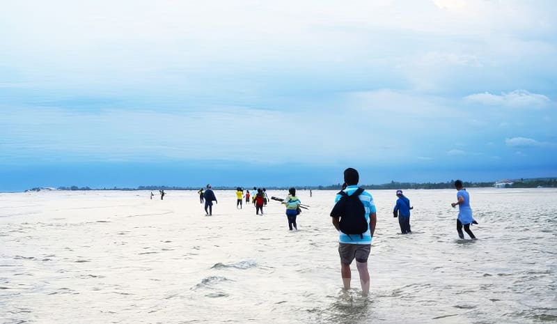 Mọi người đi bộ qua vùng nước nông trên một bãi biển rộng lớn dưới bầu trời nhiều mây, tạo nên một khung cảnh thanh bình và đầy phiêu lưu.