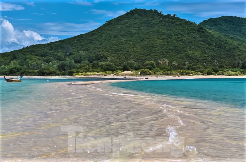 Một khung cảnh bãi biển thanh bình với con đường cát trắng dẫn đến ngọn đồi xanh mướt. Nước biển màu ngọc lam và một chiếc thuyền nhỏ càng làm tăng thêm bầu không khí yên tĩnh.