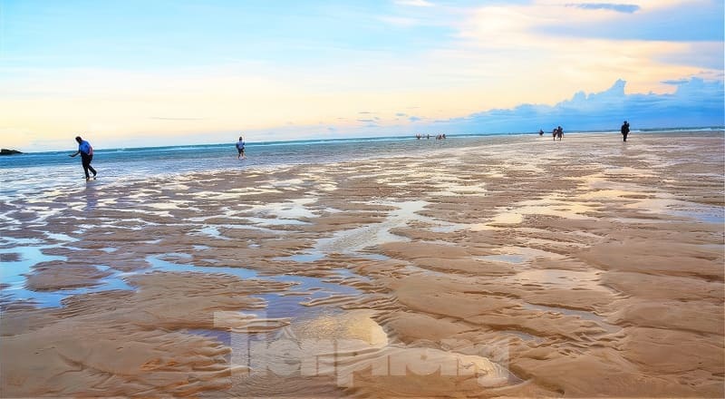 Mọi người tản bộ trên một bãi biển rộng lớn, phản chiếu ánh sáng lúc hoàng hôn, với bầu trời đầy màu sắc và đường chân trời xa xăm, tạo nên một khung cảnh thanh bình, tĩnh lặng.