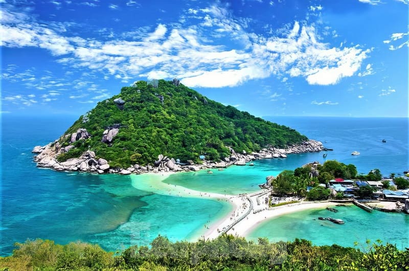 Một hòn đảo xanh tươi với dải bãi biển cát trắng mịn màng, bao quanh là làn nước màu ngọc lam dưới bầu trời xanh trong vắt điểm xuyết những đám mây rải rác.