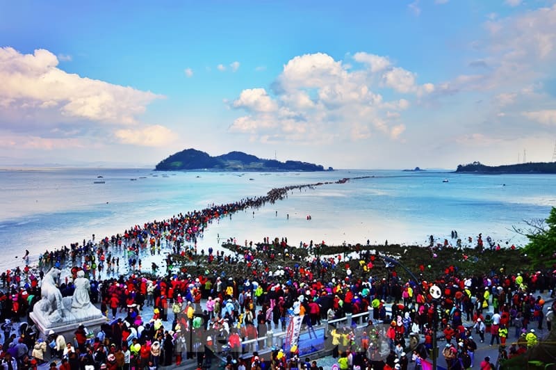 Crowds gather on a narrow path revealed by low tide, connecting to an island under a clear blue sky with scattered clouds.