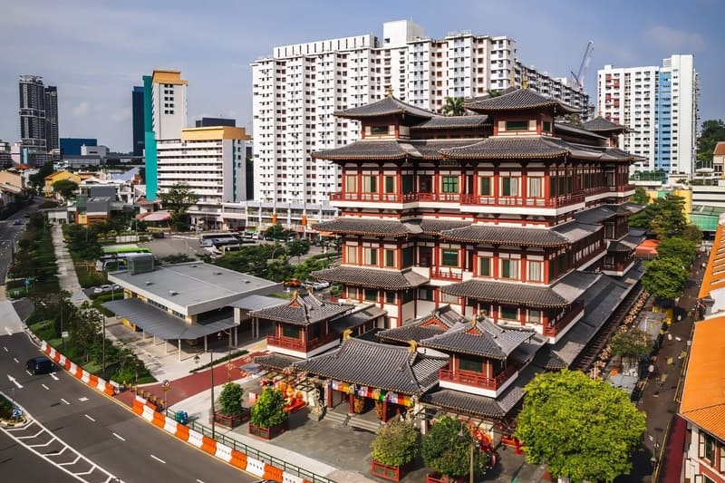 Ngôi chùa Phật giáo truyền thống nhiều tầng với những chi tiết trang trí màu đỏ và trắng cầu kỳ, nằm giữa những tòa nhà cao tầng hiện đại và một con phố đô thị nhộn nhịp.