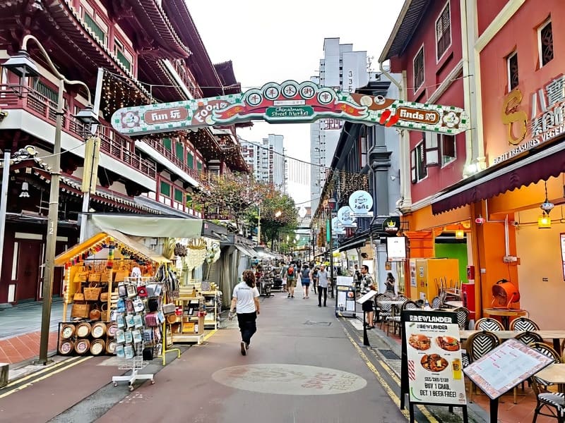 Khu chợ đường phố Chinatown nhộn nhịp với những cửa hàng đầy màu sắc, biển hiệu trang trí công phu và những người bán hàng rong sôi nổi. Mọi người tản bộ dưới một mái vòm trang trí.