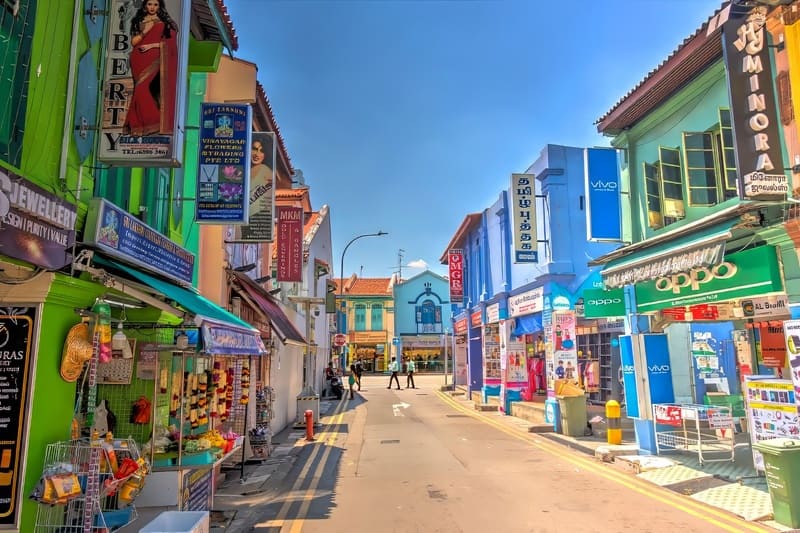 Một khung cảnh đường phố đầy màu sắc với những tòa nhà màu xanh lá cây và xanh dương rực rỡ, trưng bày vô số biển hiệu bằng nhiều ngôn ngữ khác nhau. Các cửa hàng nằm dọc theo con phố, gợi lên một bầu không khí đa văn hóa sôi động.