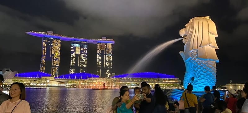 Cảnh đêm của tượng Merlion ở Singapore, phun nước, với tòa nhà Marina Bay Sands được chiếu sáng rực rỡ phía sau. Mọi người đang chụp ảnh, tạo nên một bầu không khí sống động.