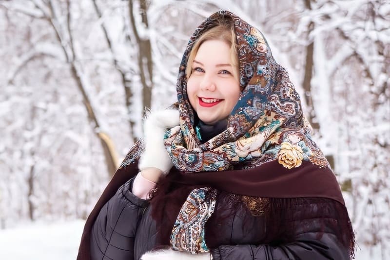 Một người tươi cười, quàng chiếc khăn nhiều màu sắc, đứng trong khu rừng phủ đầy tuyết, mặc áo khoác tối màu và đeo găng tay trắng, toát lên sự ấm áp và niềm vui trong một ngày đông.