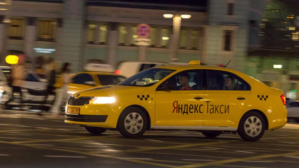 Chiếc taxi Yandex màu vàng với họa tiết ca rô chạy xuyên qua thành phố về đêm, bao quanh là những tòa nhà và người đi bộ mờ ảo, thể hiện tốc độ và sự hối hả.