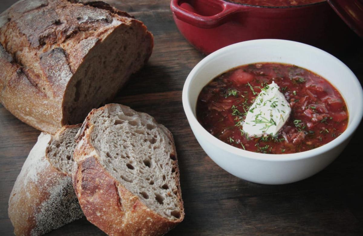 Một bát súp đỏ rưới kem chua và rau thơm, đặt cạnh những lát bánh mì nâu mộc mạc trên một chiếc bàn gỗ. Một chiếc nồi màu đỏ được nhìn thấy một phần.