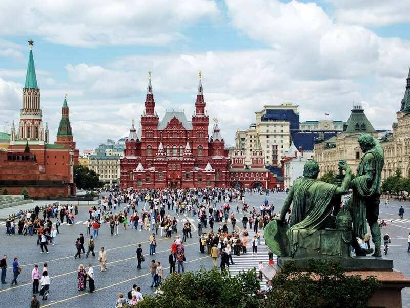 Quảng trường Đỏ rực rỡ ở Moscow; đám đông nhộn nhịp, trung tâm bảo tàng lịch sử màu đỏ, tượng màu xanh bên phải, nền trời nhiều mây vui tươi, không khí sôi động.