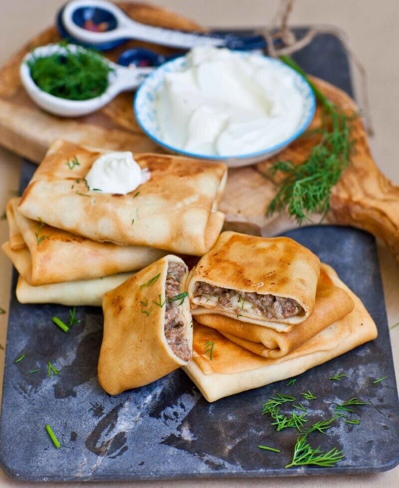 Những đĩa bánh crepe vàng óng, nhân thịt được trang trí bằng thì là tươi. Một bát kem chua và thêm thì là được đặt ở phía sau.