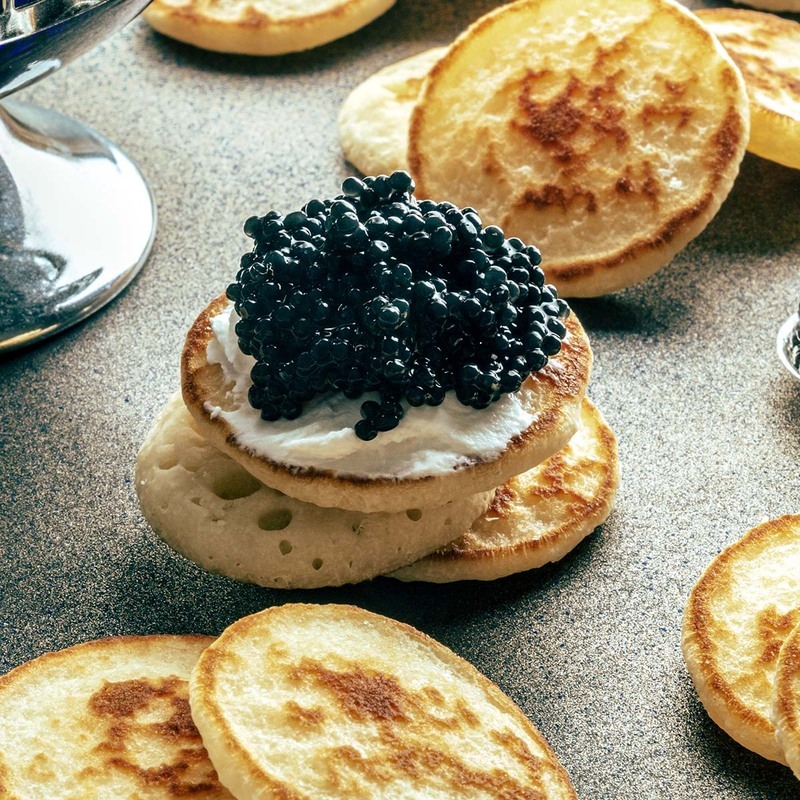 Cận cảnh bánh blinis với kem chua và trứng cá muối đen trên bề mặt màu xám sần sùi. Khung cảnh gợi lên cảm giác sang trọng và xa hoa.
