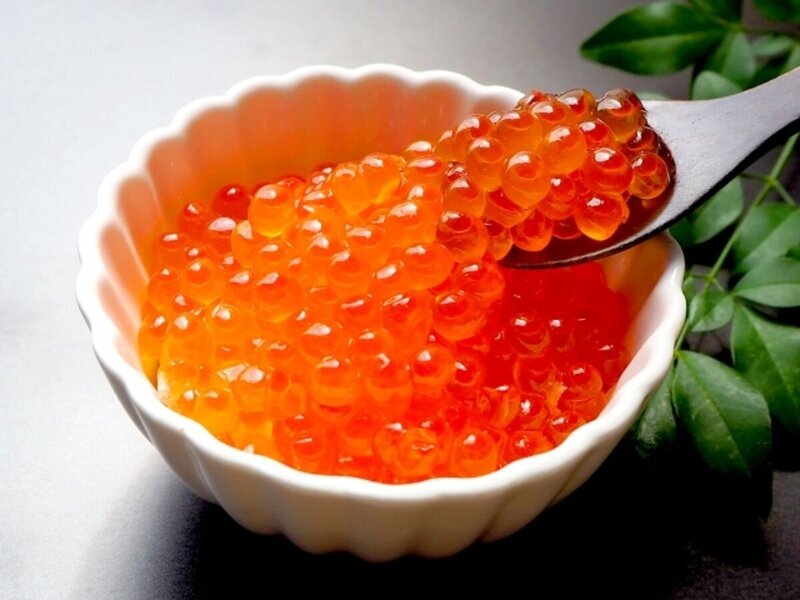 Một chiếc bát trắng đựng đầy trứng cá hồi màu cam tươi tắn, óng ánh. Một chiếc thìa múc một ít trứng cá, bên cạnh là những chiếc lá xanh tươi, tạo nên một cảnh tượng tươi mới và trang nhã.