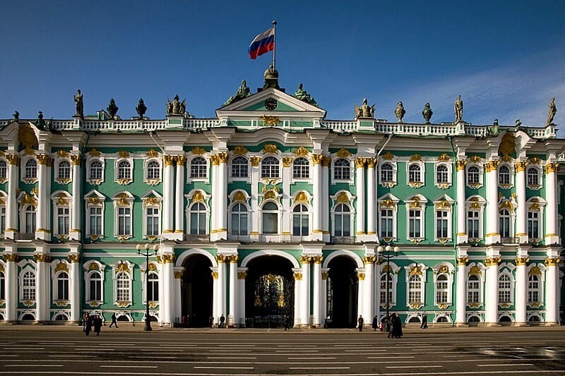 Hình ảnh cho thấy Cung điện Mùa đông ở St. Petersburg, nổi bật trên nền trời xanh. Mặt tiền trang trí công phu có màu xanh lá cây và trắng với các chi tiết mạ vàng, trên đỉnh là quốc kỳ Nga.