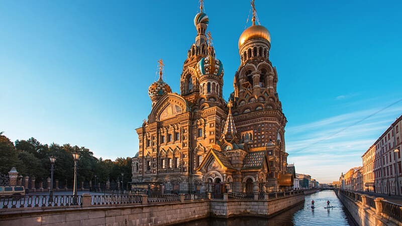 Hình ảnh mô tả Nhà thờ Chúa Cứu Thế trên Máu Đổ ở St. Petersburg, Nga, với những mái vòm rực rỡ được chiếu sáng bởi ánh nắng vàng, nằm bên cạnh một con kênh yên tĩnh.