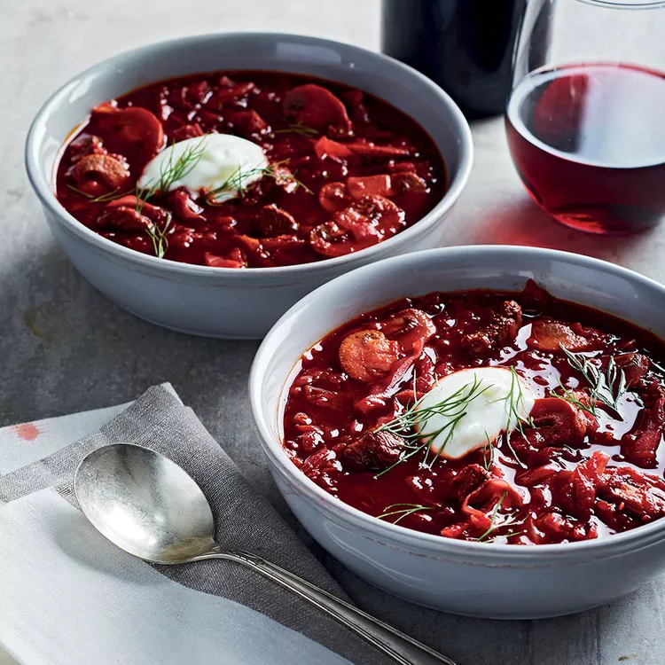 Hai bát súp borscht đỏ tươi, chan thêm kem chua và thì là tươi, được đặt trên bàn, bên cạnh một ly rượu vang và một chiếc thìa.