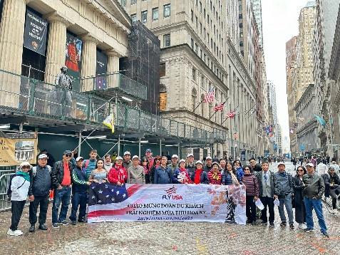 Một nhóm người đứng trên đường phố, cầm một tấm biểu ngữ lớn được trang trí công phu. Khung cảnh được bao quanh bởi những tòa nhà cao tầng, và một số lá cờ có thể nhìn thấy. Không khí rất náo nhiệt và vui tươi.