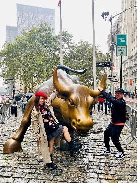 Một người phụ nữ đội mũ nồi đỏ và một người đàn ông đội mũ đen tạo dáng vui vẻ bên bức tượng Bò tót đang lao tới ở thành phố New York, trên một con phố lát đá cuội.