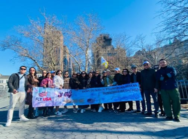 Một nhóm người đứng thành hàng ngoài trời vào một ngày nắng đẹp, tay cầm một biểu ngữ. Những hàng cây trơ trụi lá và những tòa nhà cao tầng hiện lên ở phía xa.