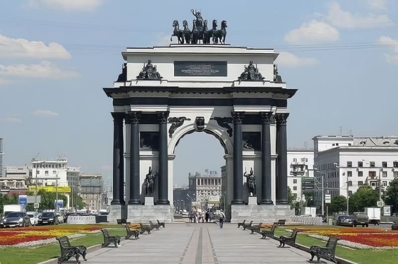 Cổng vòm chiến thắng trong khung cảnh thành phố với những bức tượng trên đỉnh, bao quanh là những luống hoa, ghế đá và các tòa nhà liền kề. Bầu trời trong xanh.