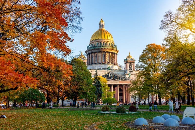 Cảnh mùa thu tại nhà thờ St. Isaac với mái vòm vàng rực rỡ, bao quanh là những tán cây xanh và cam rực rỡ, cùng những người đang tản bộ trong công viên ngập nắng.